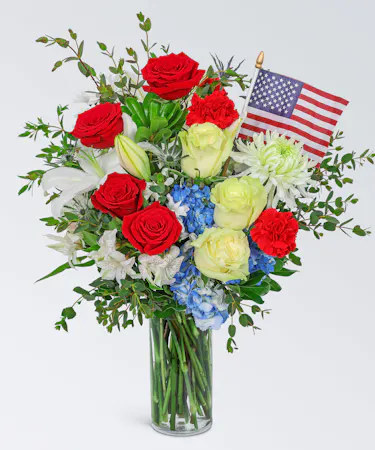 A vibrant red, white, and blue color scheme, and includes gorgeous hydrangea, roses, carnations, lilies, mums, alstroemeria, and other premium foliage in a vase