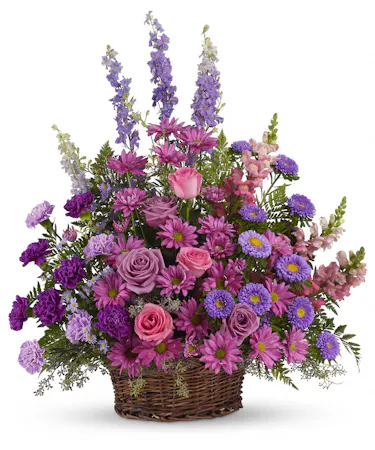A profusion of purple, pink and lavender blooms such as roses, carnations, larkspur, and much more are delivered in a round wicker basket. 
