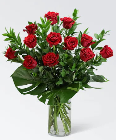 One dozen red roses with premium greenery and monstera leaf in a clear glass vase