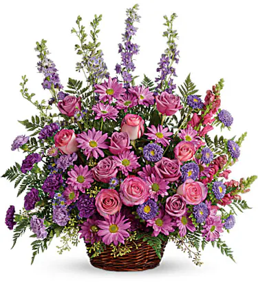 A profusion of purple, pink and lavender blooms such as roses, carnations, larkspur, and much more are delivered in a round wicker basket. 