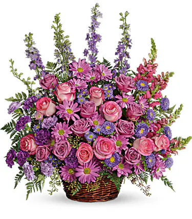 A profusion of purple, pink and lavender blooms such as roses, carnations, larkspur, and much more are delivered in a round wicker basket. 
