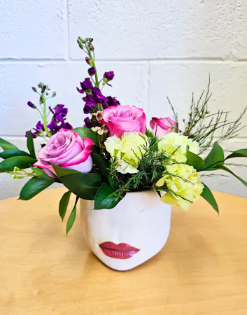 White ceramic container with red lips filled with lavender roses, purple stock, green carnations and lush greenery