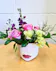 White ceramic container with red lips filled with lavender roses, purple stock, green carnations and lush greenery