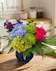 A blue hued glass vase with purple stock, blue and green hydrangea, hot pink roses and carnations and lush greenery