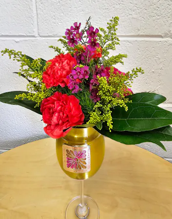 Hot pink carnations, orange spray roses, accent flowers and greenery in a Queen's Jewels embellished wine glass