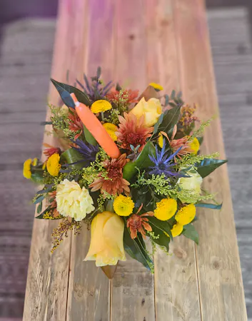 a single taper candle nestled in a Thanksgiving arrangement of orange carnations, fall chrysanthemums, green button mums and fall foliage and accents