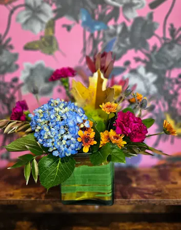 A leaf wrapped glass cube with blue hydrangea, purple carnations, eryngium, sunset leucadendron and greenery
