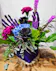 black roses, blue hydrangea, purple carnations, lots of greenery in a purple cube with purple velvet skeleton hands