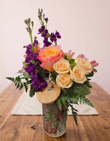 A vibrant Free Spirit rose, peach spray roses and purple stock arrangement in a reusable patterned tumbler with a bamboo lid