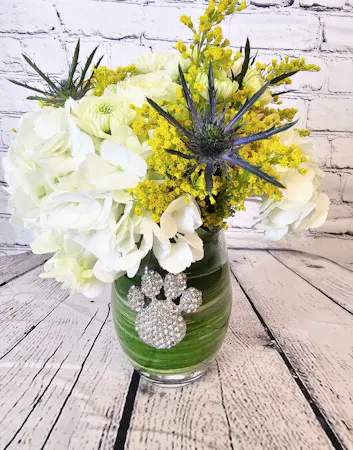 White hydrangea, roses, chrysanthemums are accented with blue eryngium and yellow solidago. Tucked into our Queen's Jewels winegass embellished with a rhinestone paw print