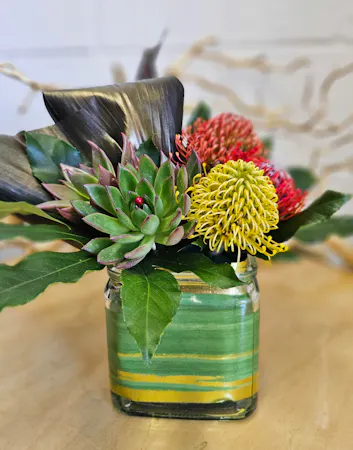 3 pincushion protea, 2 succulents, tropical foliage, a gold leaf-lined cube, and black ti leaf in a modern style design