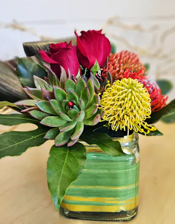 3 pincushion protea, 2 succulents, tropical foliage, a gold leaf-lined cube, and black ti leaf in a modern style design