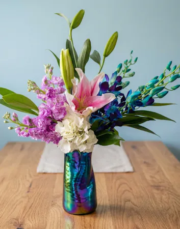 A stunning floral arrangement featuring pink Stargazer lilies, exotic blue orchids, and white hydrangeas in an iridescent vase.