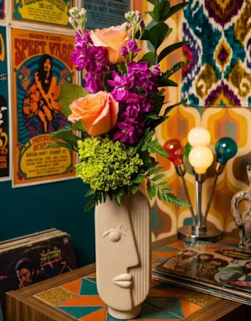 . This unique sculptural face vase overflows with vibrant coral roses, purple stock, and neon greens—the perfect gift for dreamers and design lovers.