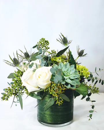 A compact arrangement of white roses, succulent, eryngium, green hydrangea, eucalyptus are arranged in a cylinder vase with a leaf wrap. 